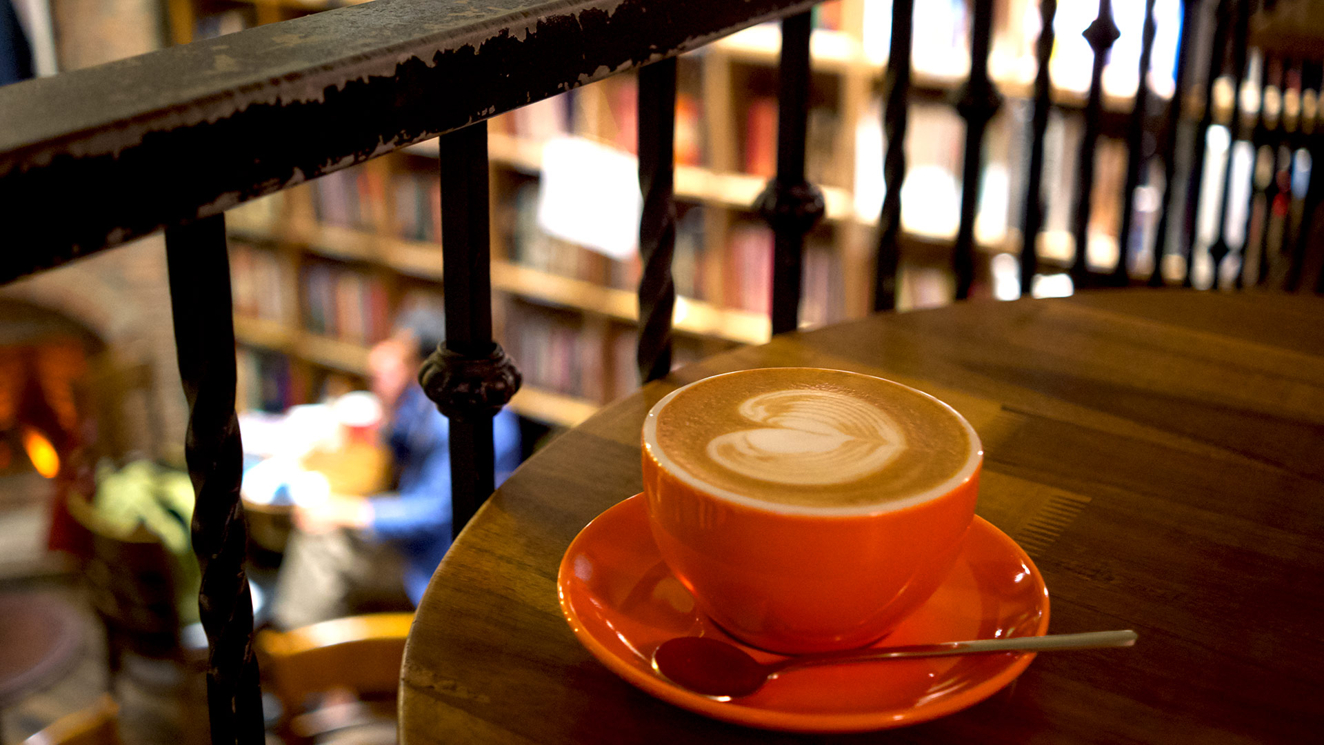A single orange cup of coffee on a wooden table on the mezzanine of a bookshop café in Hanoi, wrought-iron railing in the foreground and shelves of books going soft behind.