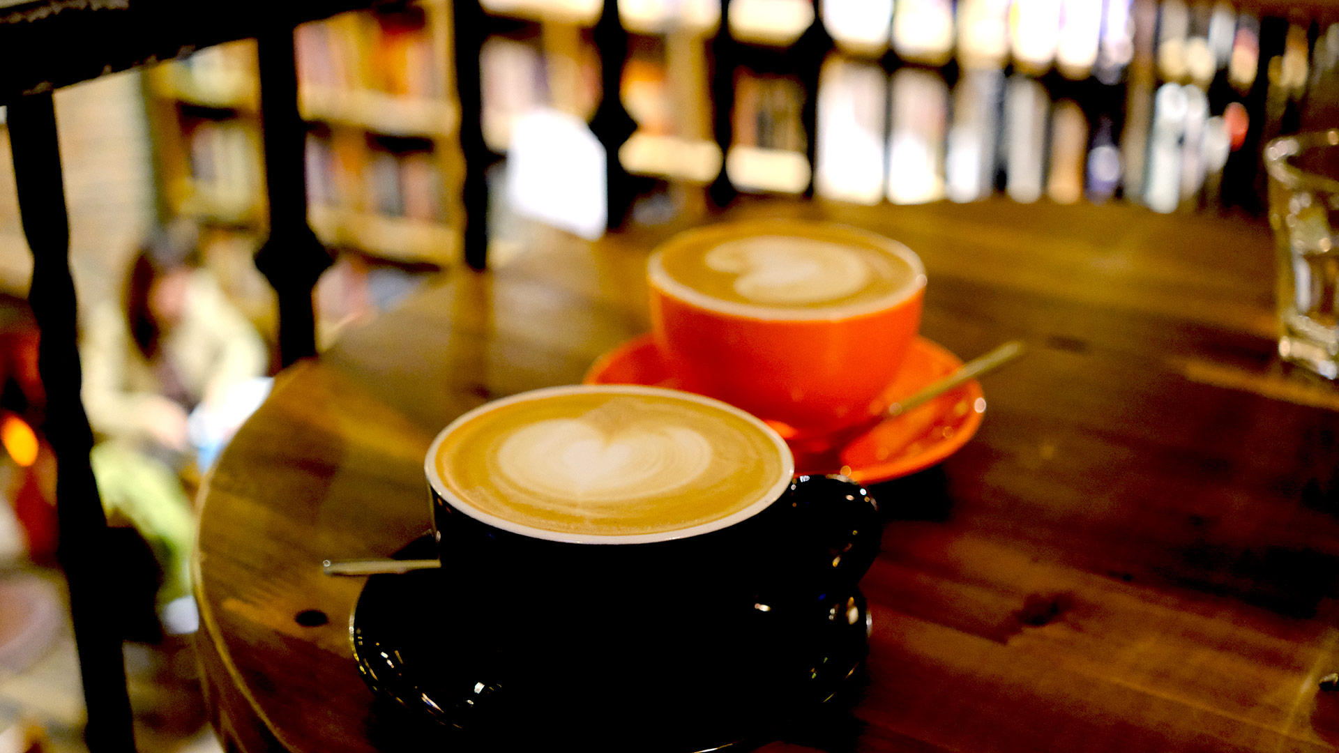 Two cups of coffee on a wooden table in a Hanoi bookshop café, one black cup in the foreground and one orange behind, shelves of books out of focus beyond.