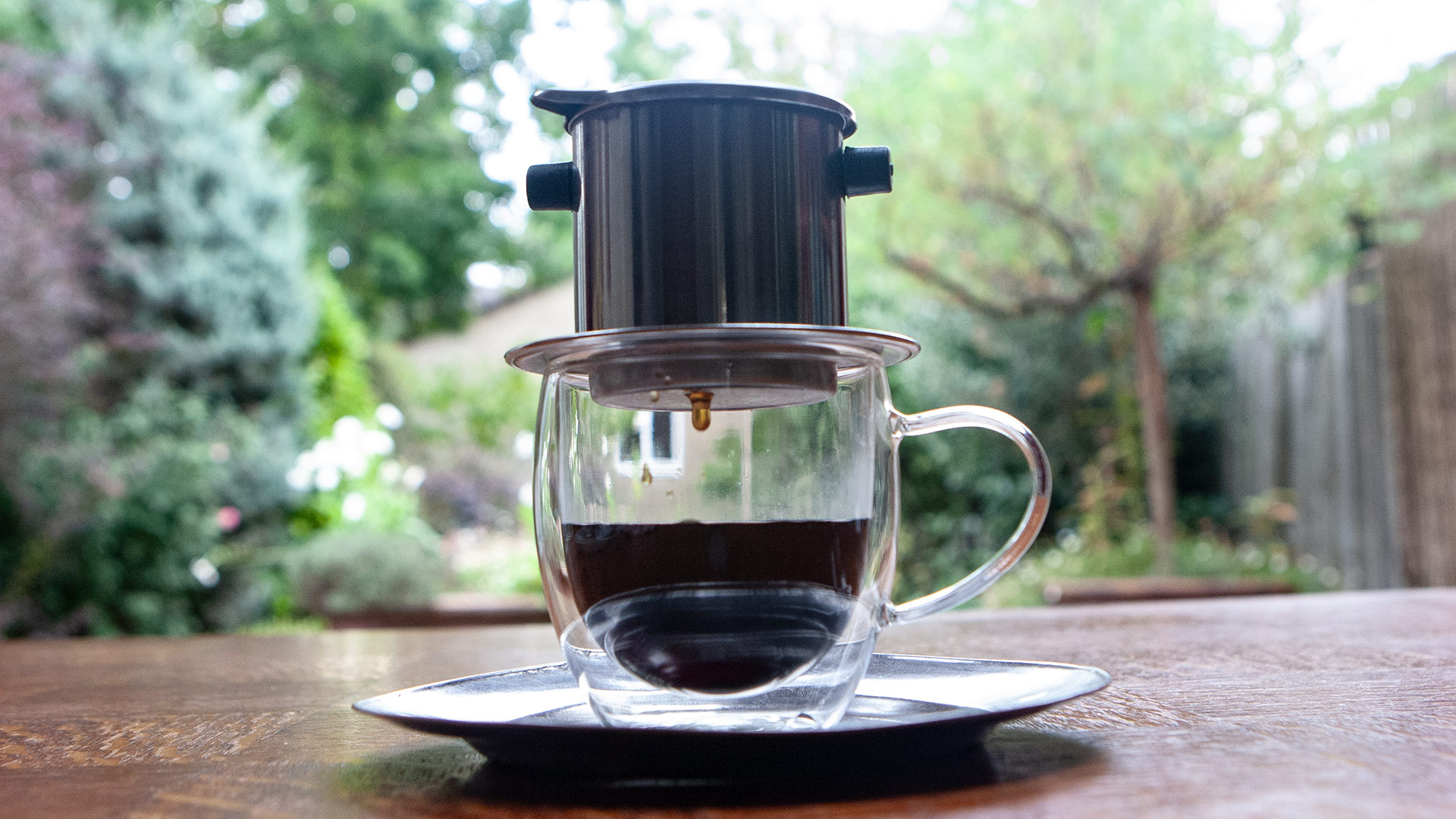 A metal phin filter sits on a glass cup on a garden table, a single drop of coffee falling from its base into the dark coffee below.